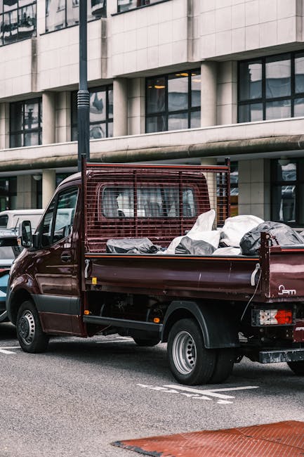 A small burgundy flatbed truck parked on a city street next to a modern office building with large glass windows and a concrete facade. The truck's flatbed is loaded with various bags and and loose ru