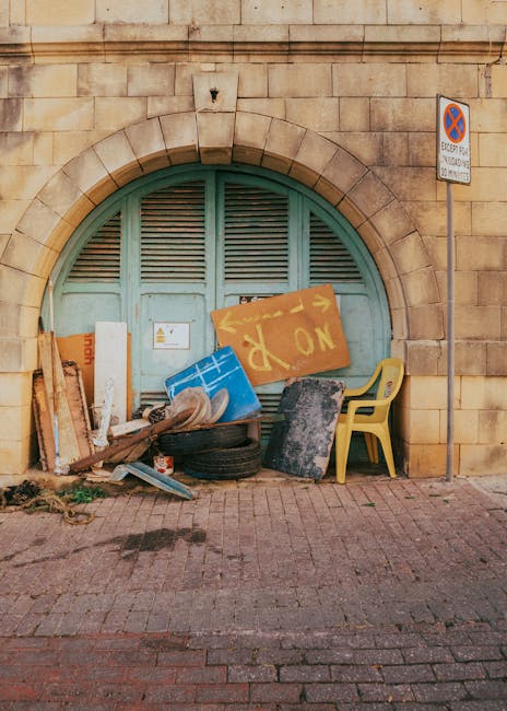 A small burgundy flatbed truck parked on a city street next to a modern office building with large glass windows and a concrete facade. The truck's flatbed is loaded with various bags and and loose ru