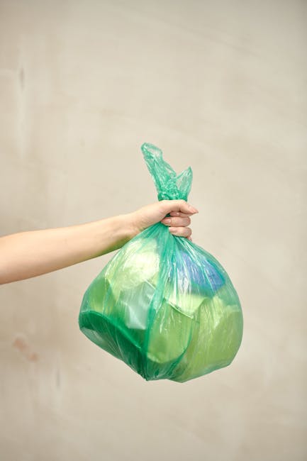 Close-up of a person wearing a multicolored checkered shirt, blue jeans, and bright green gloves, holding open a black plastic rubbish bag with both hands, positioned outdoors on a grassy lawn. The ba