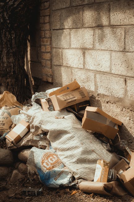 A collection of mixed waste and rubbish piled on the ground in an outdoor setting, featuring black and transparent plastic bags filled with refuse, a large black rubber tire leaning against the pile,